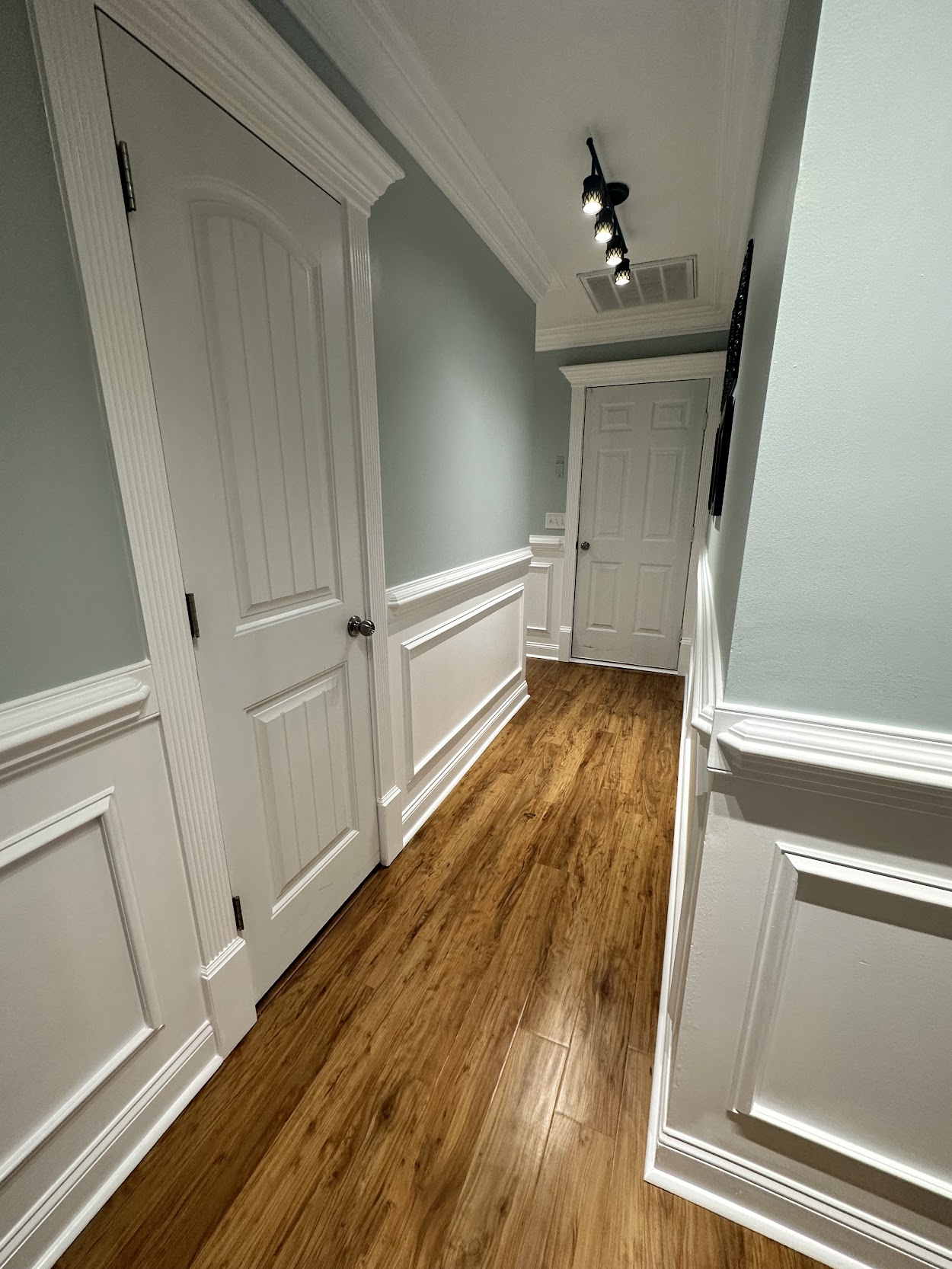Hallway with warm wood-look flooring, white wainscoting, crown molding, and two white doors